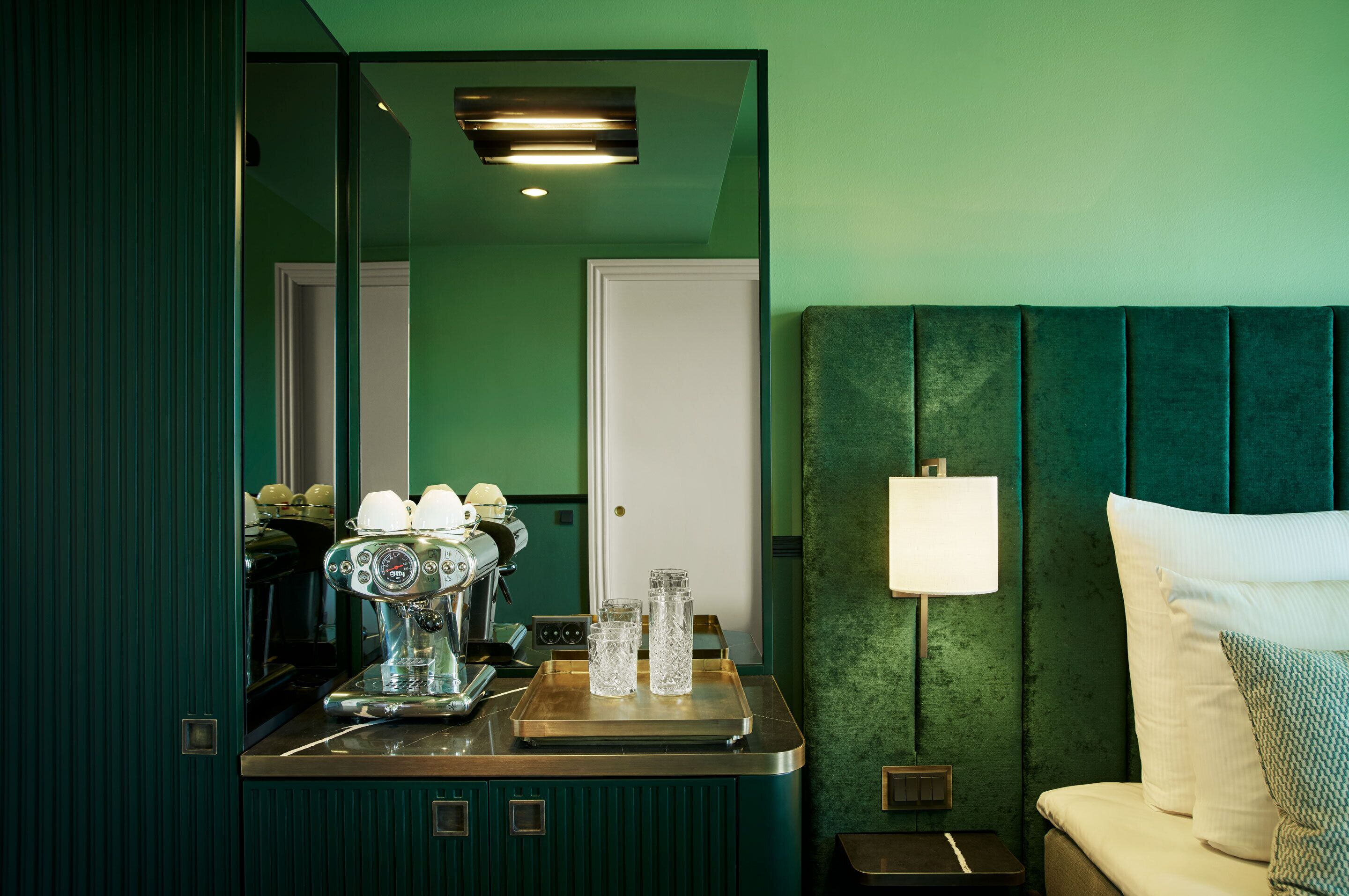 Una habitación con pared verde, cama con cabecera verde, mesita de noche con lámpara y una cafetera sobre una bandeja.