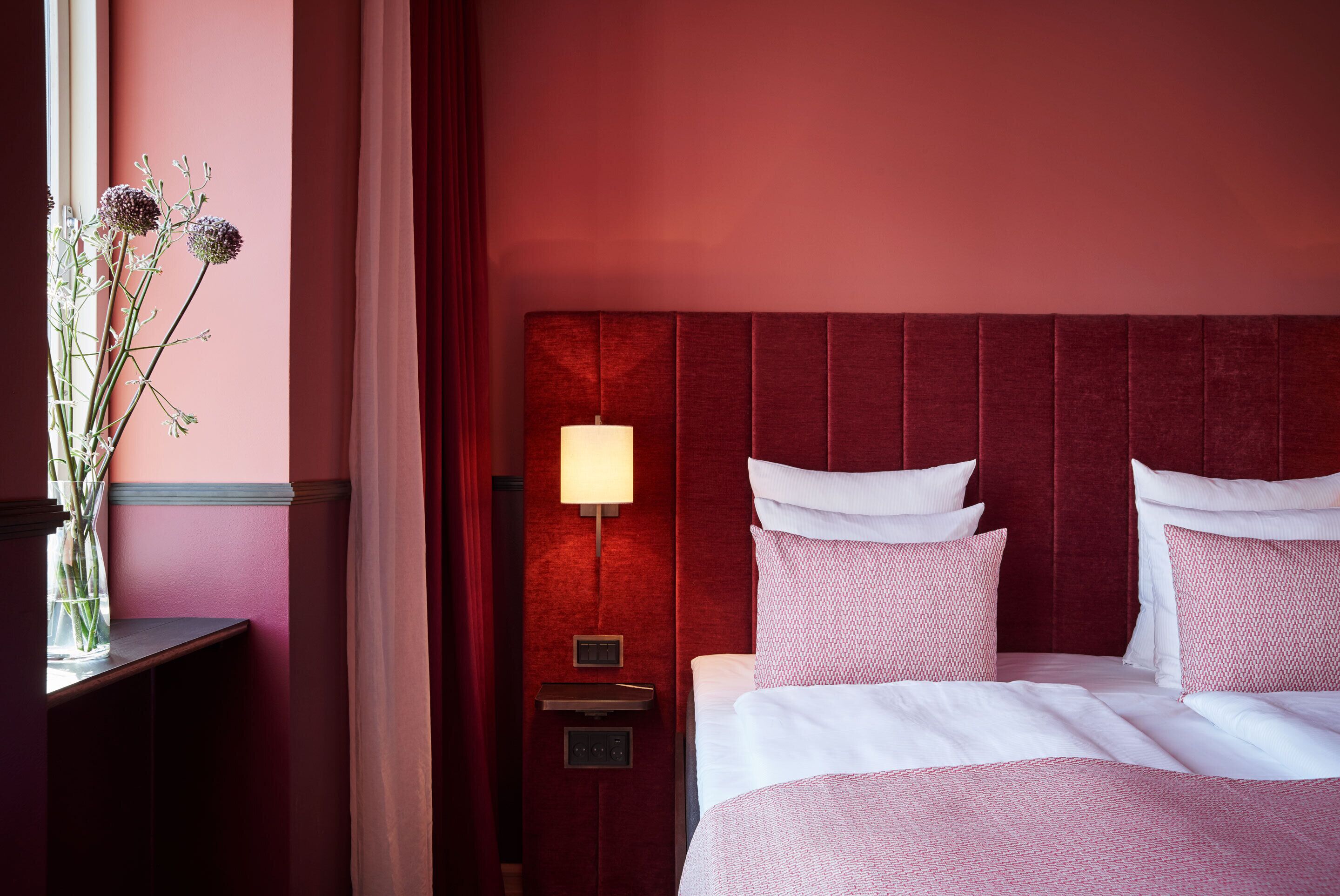 Una habitación de hotel con cabecera roja, una mesita de noche con lámpara y una cama con ropa de cama blanca y rosa.