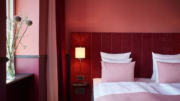 Una habitación de hotel con cabecera roja, una mesita de noche con lámpara y una cama con ropa de cama blanca y rosa.