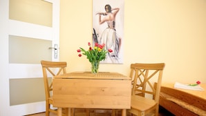 Una habitación con una mesa y sillas de madera, un florero con flores rojas y amarillas, un cuadro en la pared y una cama con cubre cama blanco.