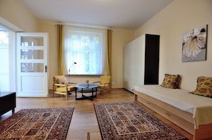 Una habitación con una cama, dos sillas, una mesa, un ventanal con cortinas y un cuadro en la pared.