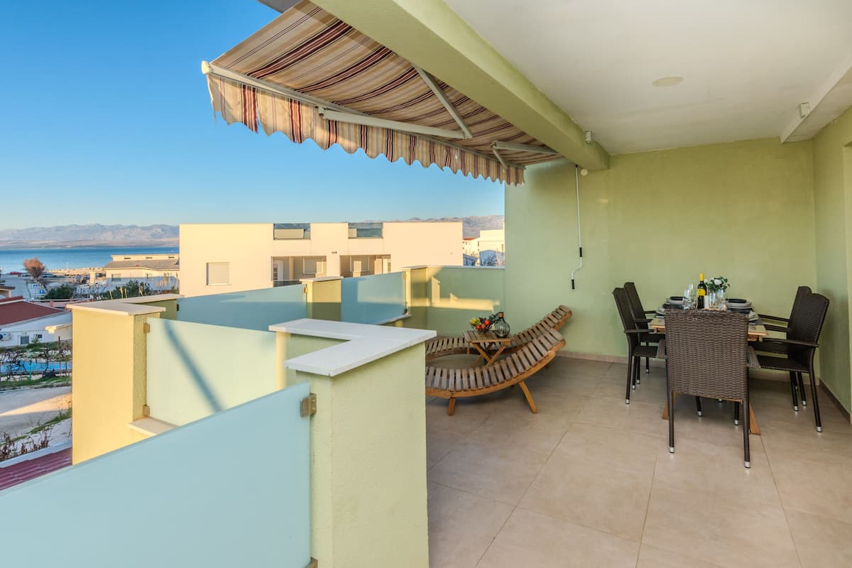 Ein Balkon mit Meerblick, ein Essbereich mit Stühlen und einem Tisch sowie eine überdachte Sitzgruppe mit gestreifter Markise.