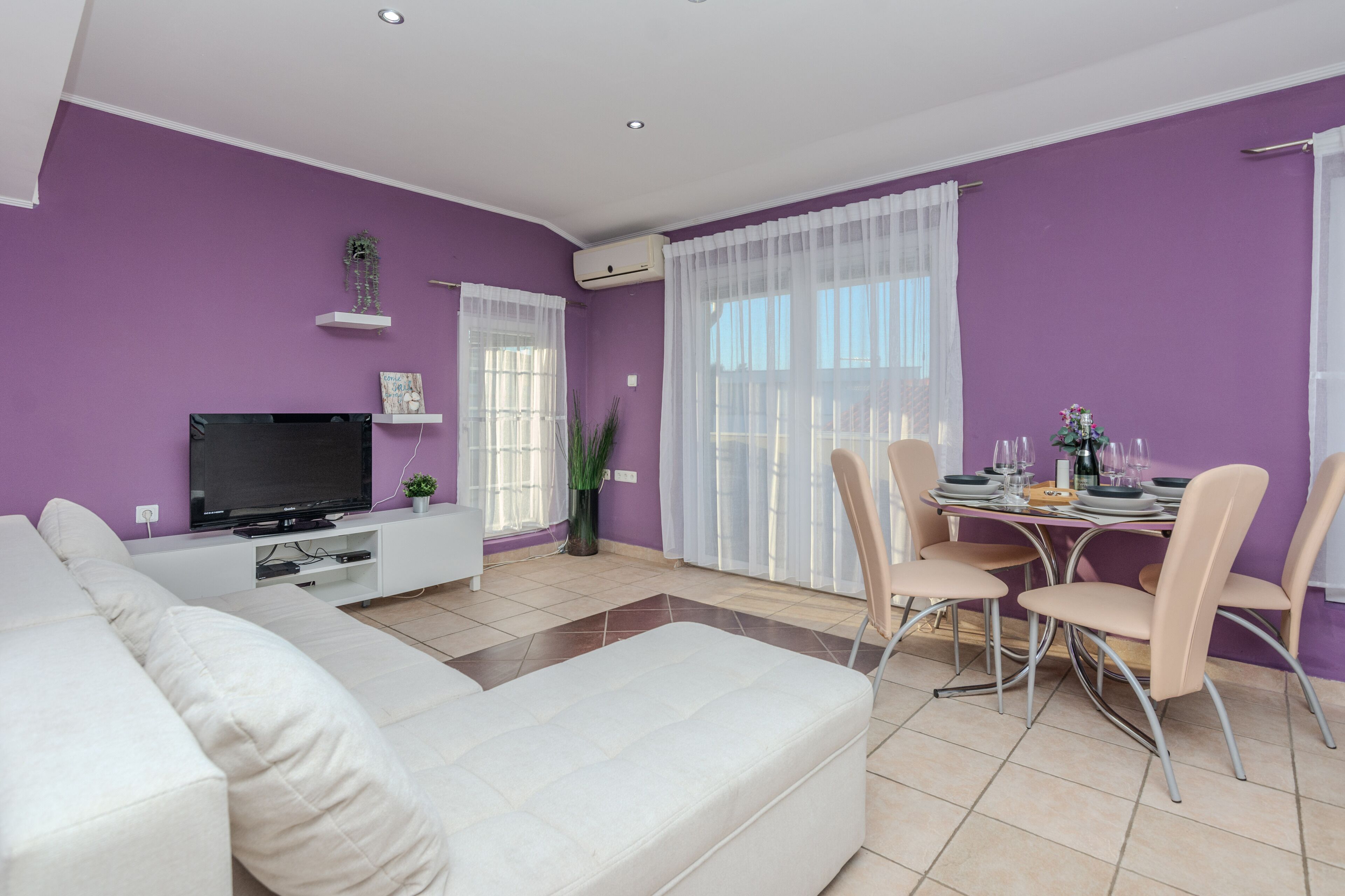 A modern living room with a white sofa, a flat-screen TV, a dining table with chairs, and a purple wall.