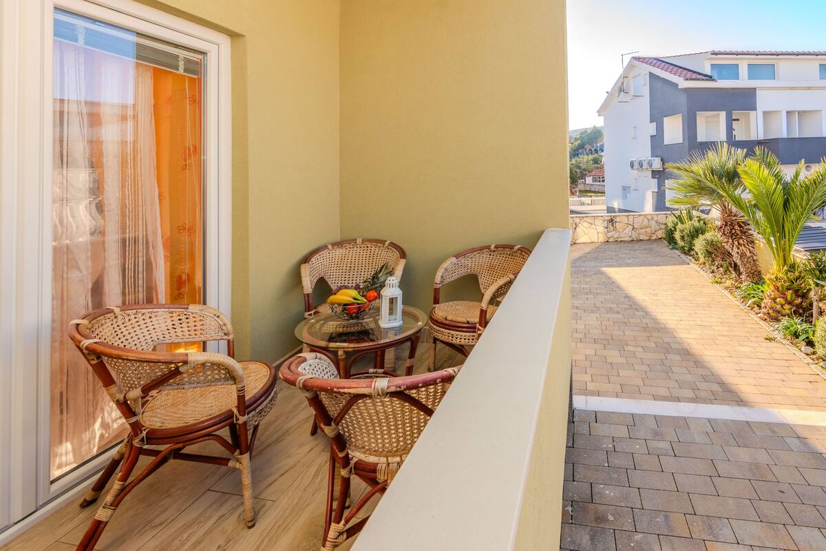 Ein Balkon mit Korbstühlen, einem Tisch mit Obst und einer Laterne, und Blick auf ein Gebäude mit Ziegeldach.
