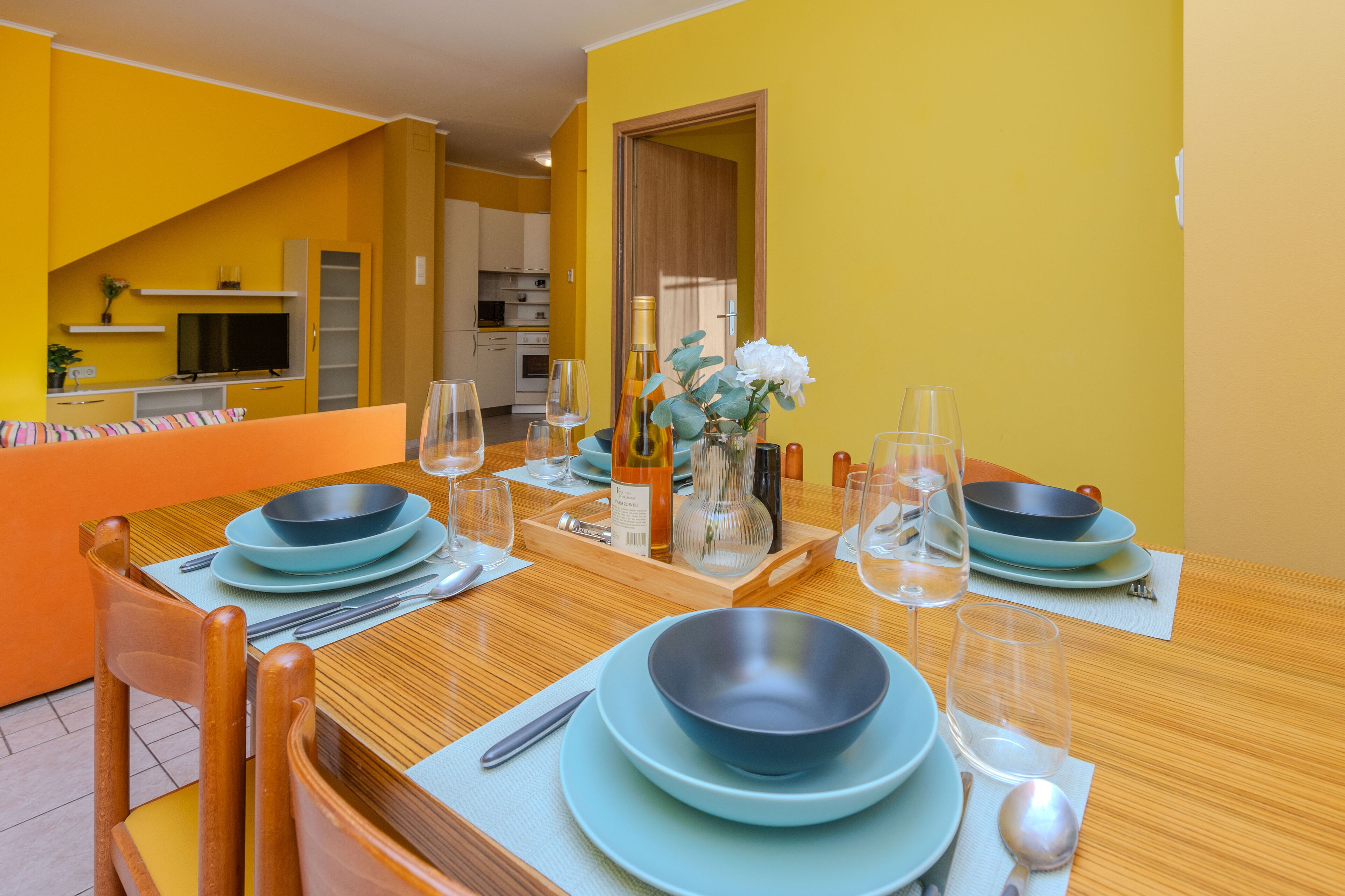 A dining area with a wooden table set for four, surrounded by yellow walls and a kitchen in the background.