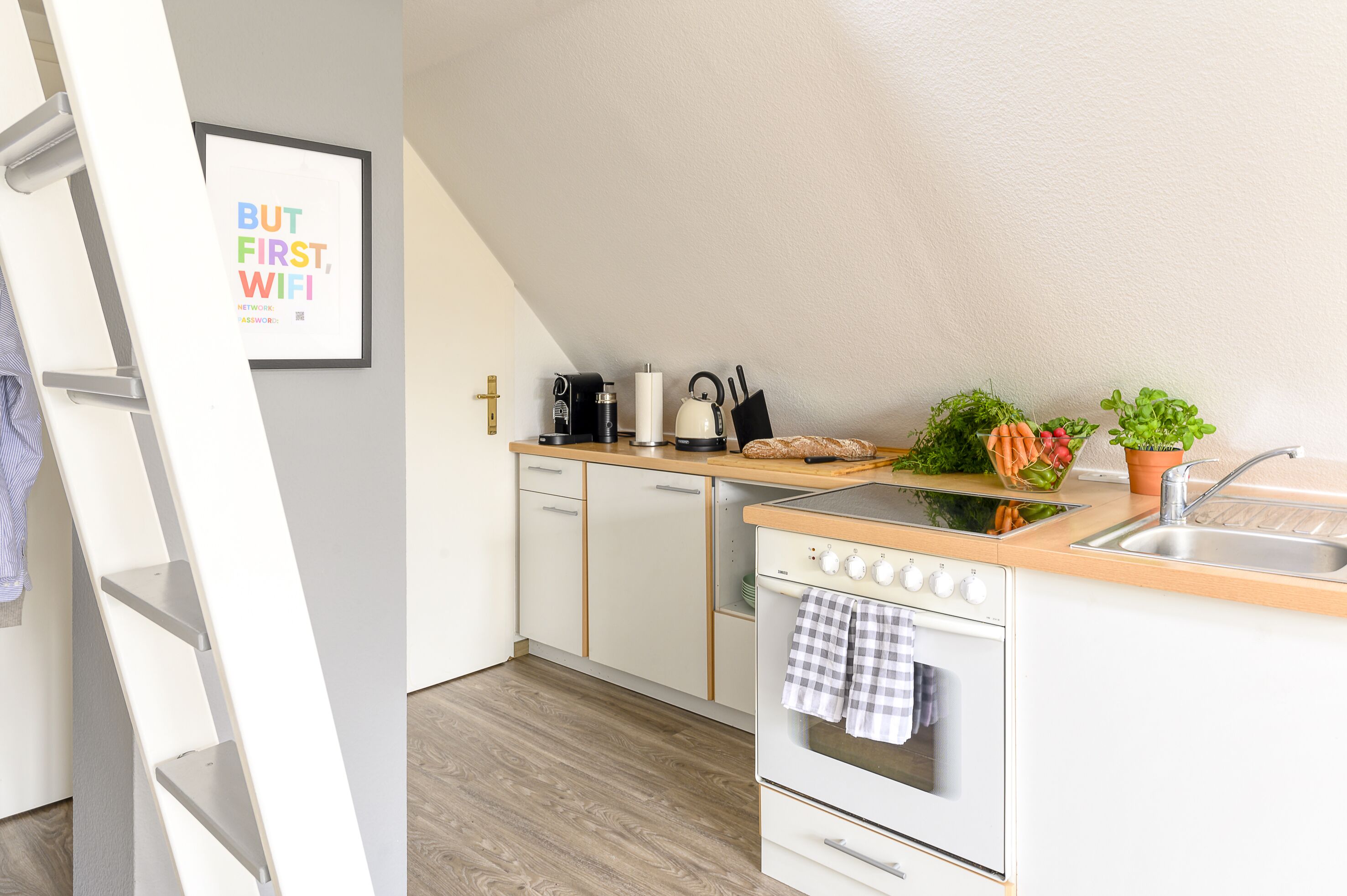 A modern kitchen with white cabinets, a wooden countertop, a stove, and a sink. There is a framed picture on the wall and a ladder leaning against the wall.