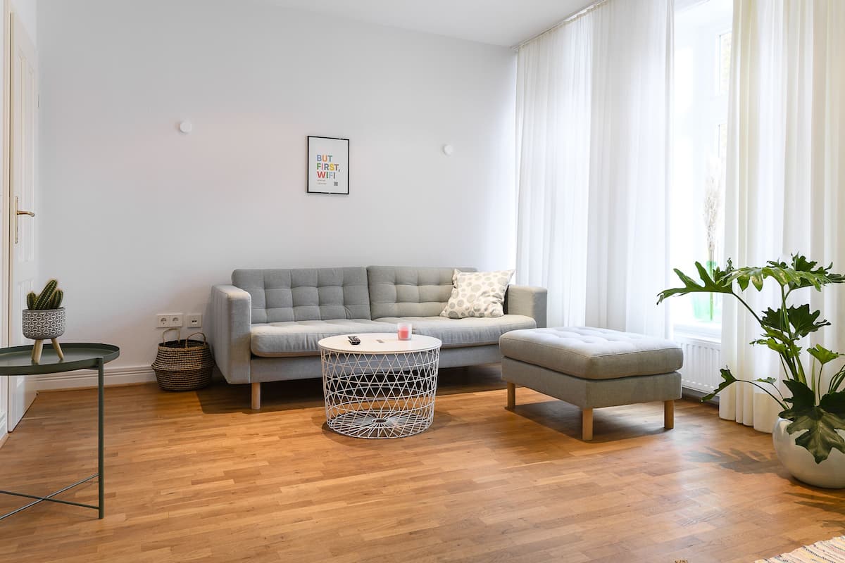 Una sala moderna con un sofá gris, una mesita redonda y un otoman gris. Hay una planta en maceta a la derecha y una pequeña mesa con un cactus a la izquierda.