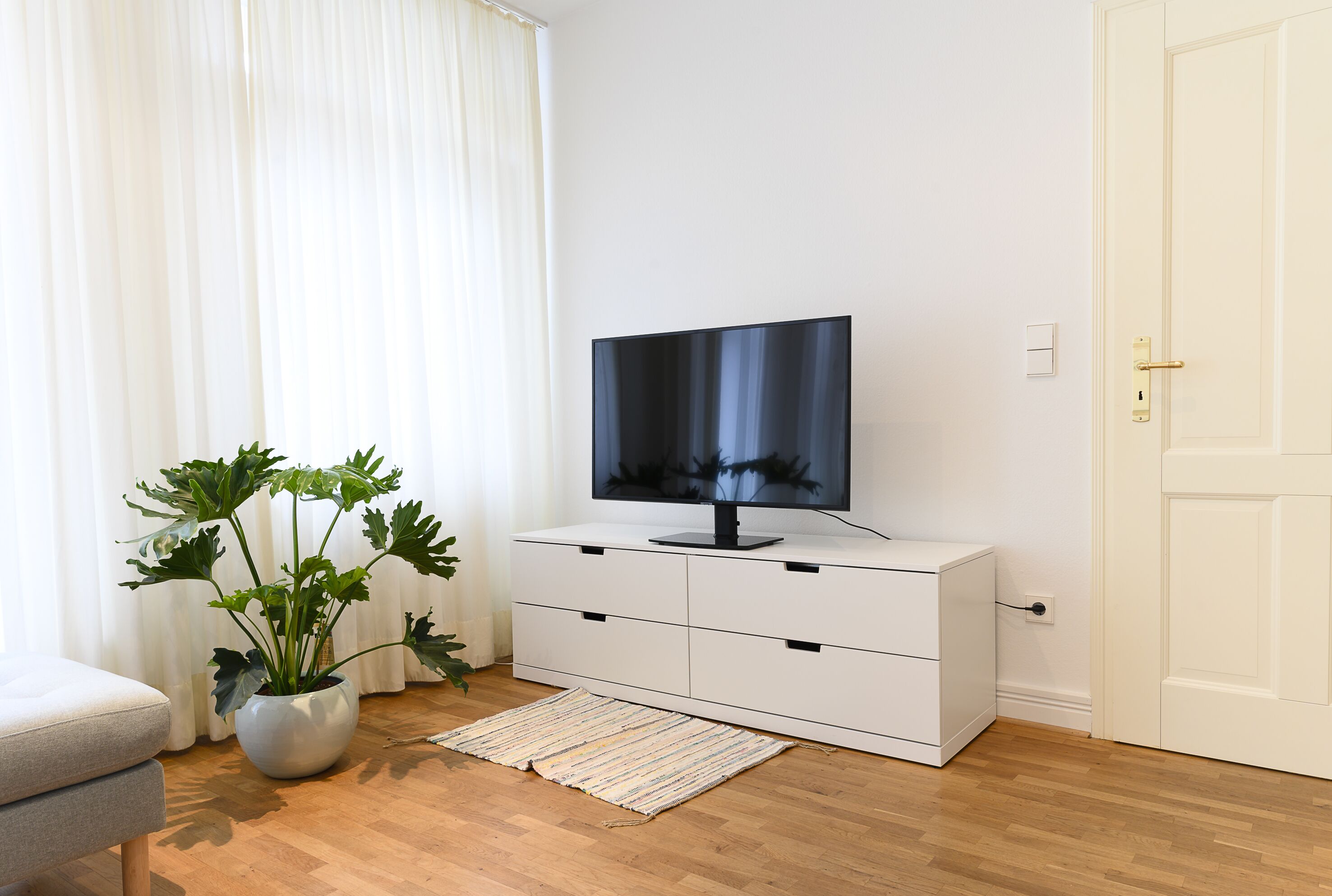 Ett modernt vardagsrum med en platt-TV placerad på ett vitt skåp, en krukväxt och ett trägolv.