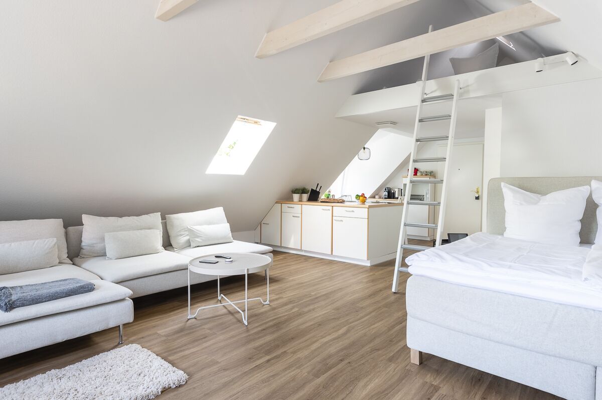 Ein moderner, gut beleuchteter Raum mit Schlafgalerie, einem weißen Sofa, einem Bett, einem kleinen runden Tisch und einer Küchenzeile mit Schränken und einer Leitern.