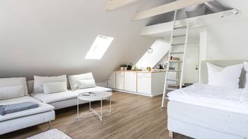 Ein moderner, gut beleuchteter Raum mit Schlafgalerie, einem weißen Sofa, einem Bett, einem kleinen runden Tisch und einer Küchenzeile mit Schränken und einer Leitern.
