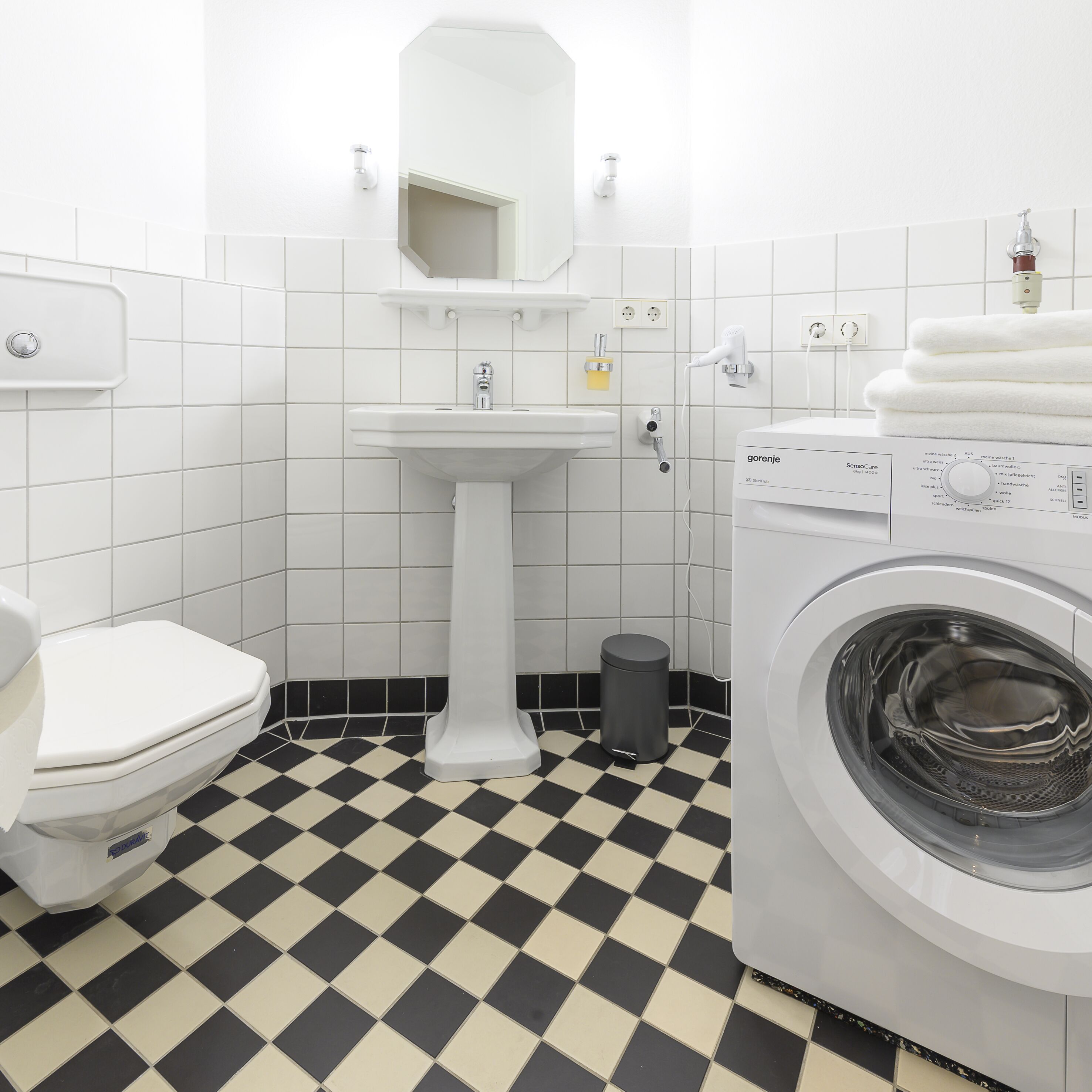A bathroom with a white sink, toilet, and washing machine, featuring a black and white checkered floor.