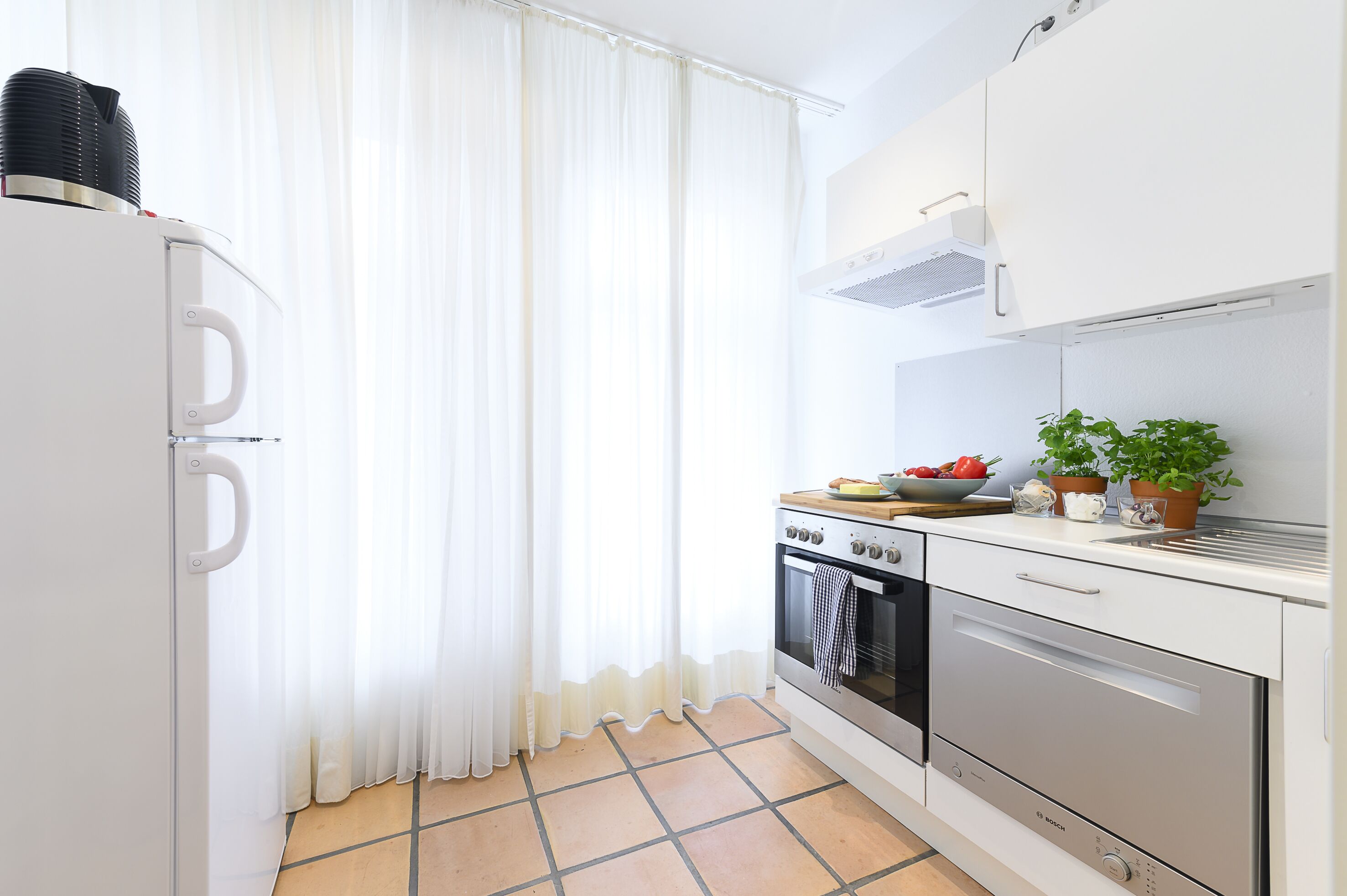 A compact kitchen with white cabinets, a stove, oven, and a sink. There is a window with sheer curtains and a tiled floor.