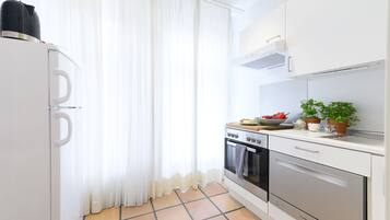 A compact kitchen with white cabinets, a stove, oven, and a sink. There is a window with sheer curtains and a tiled floor.