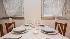Una mesa de comedor dispuesta con platos blancos, vasos y cubiertos, con una vela encendida en el centro, cortinas translúcidas y una ventana al fondo.