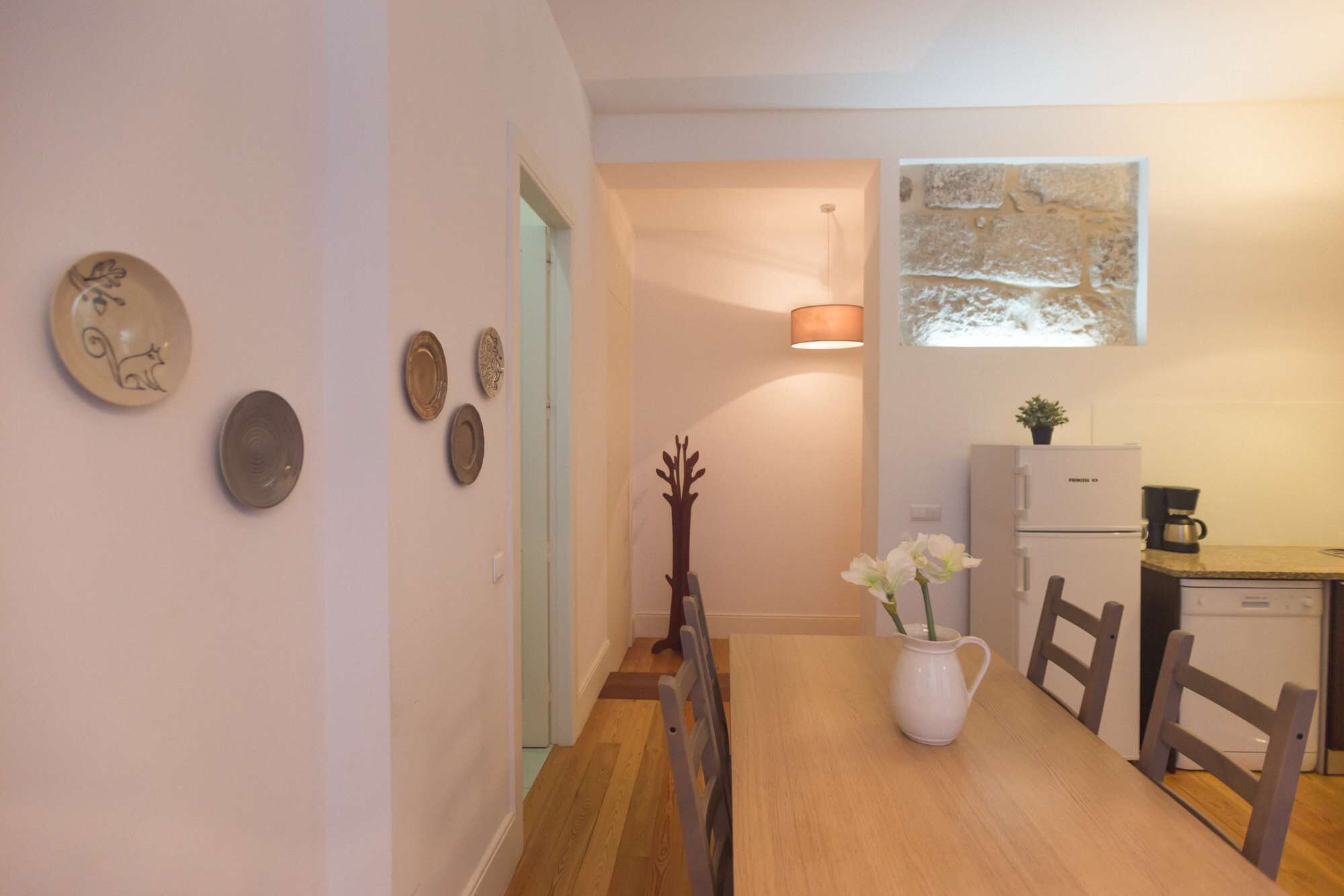 A dining area with a wooden table, chairs, a white pitcher with flowers, and a refrigerator.