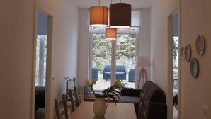 A dining area with a long table, chairs, hanging pendant lights, and a view of outdoor seating through a glass door. - Two-Bedroom Apartment (Porto)