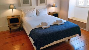 Una cama bien hecha con sábanas blancas y una manta azul, flanqueada por dos mesitas de noche con lámparas, y dos obras de arte enmarcadas en la pared.