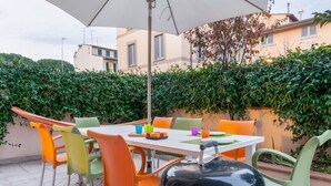 An outdoor patio with a table and chairs, a grill, and a white umbrella. - GuestHost - Comfortable 50sqm apartment with tiled patio that can accommodate up to 4 people. Located on the ground floor of a modern building of recent construction, the apartment is equipped with private parking space for a motorbike (inside the patio) (Firenze)