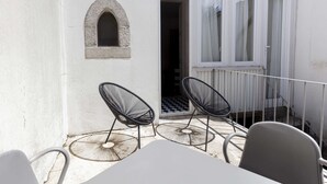 Un petit espace extérieur aménagé avec deux chaises en métal et une table, un bâtiment blanc doté d’une petite fenêtre en arc de cercle et une porte donnant sur un balcon abrité.