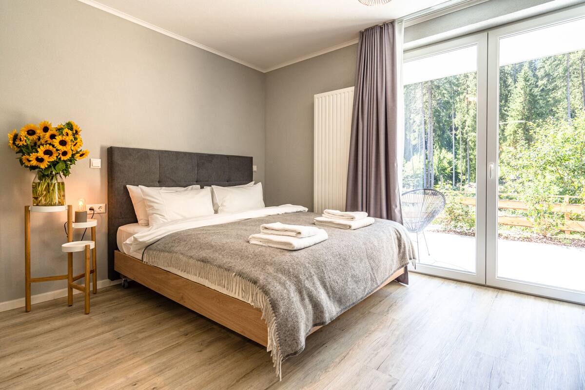Ein modernes Schlafzimmer mit einem großen Bett, einem Nachttisch mit einer Vase Sonnenblumen und Blick auf einen Garten durch eine Glastür.