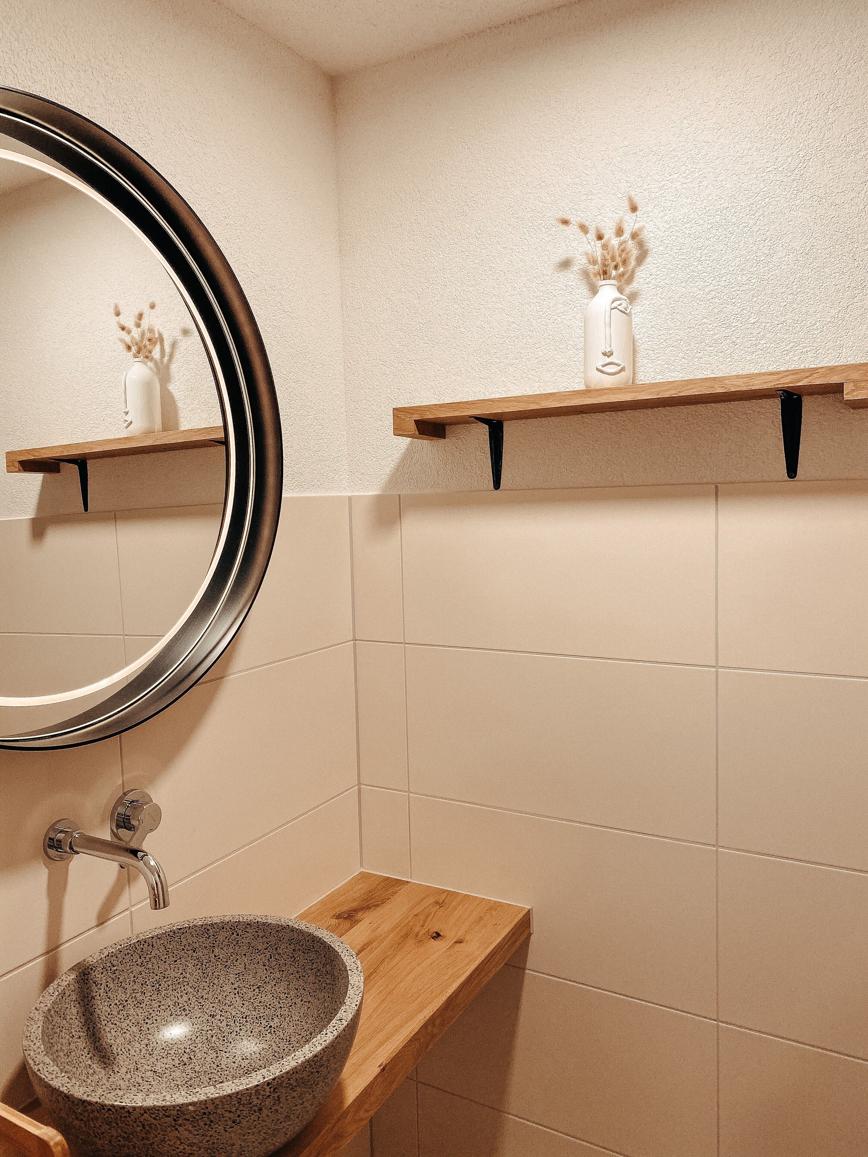 Un baño con un espejo redondo, un estante de madera con un jarrón y un lavabo de piedra.