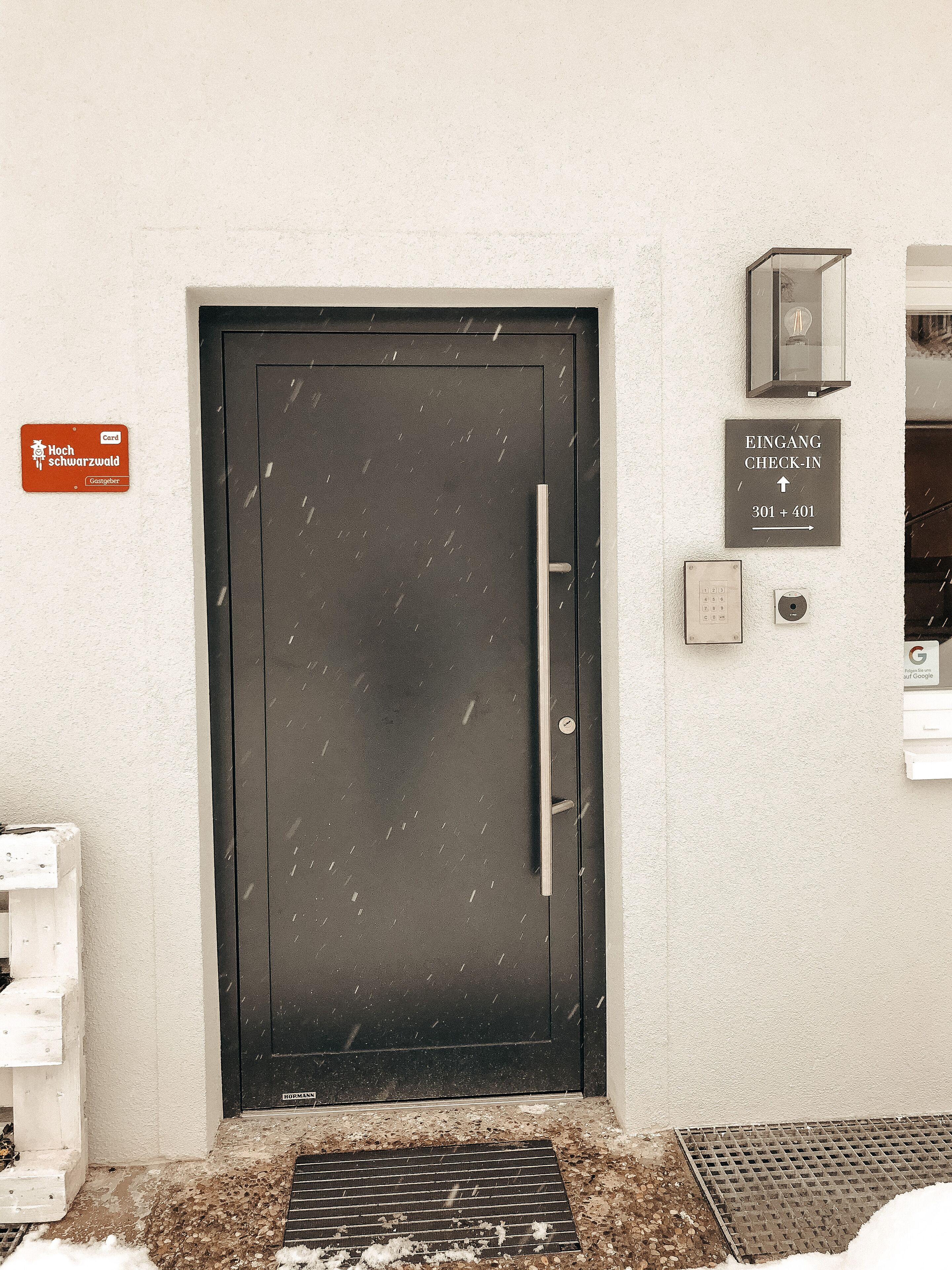 L’entrée d’un hôtel, avec une porte noire, une enseigne indiquant « Entrée – Enregistrement » et une autre mentionnant « Haute sécurité ».