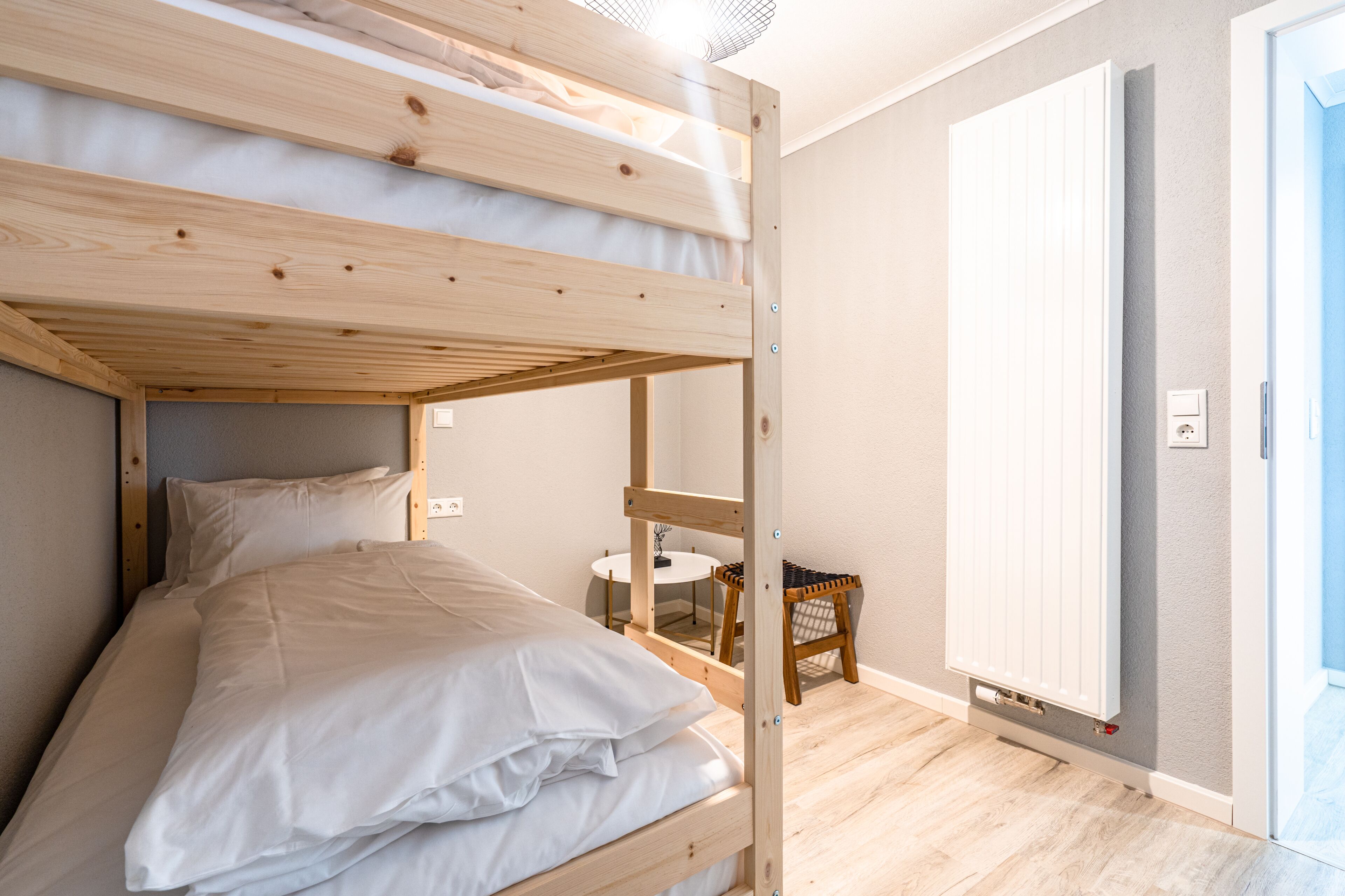 A bunk bed with white bedding, a small round table, and a chair in a room with wooden flooring and a white door.