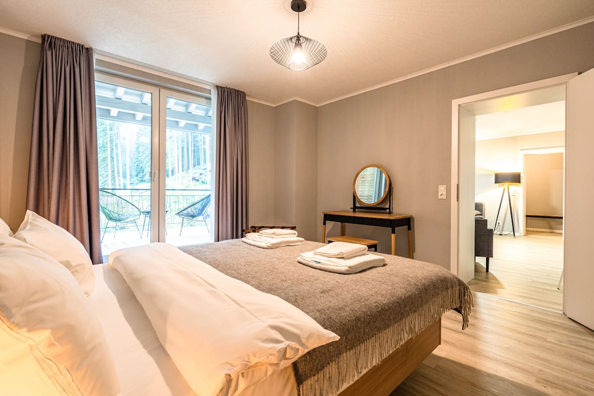 Ein Hotelzimmer mit einem Bett, einem Nachttisch mit Lampe, einem Spiegel und Blick ins Freie durch ein Fenster mit Vorhängen.