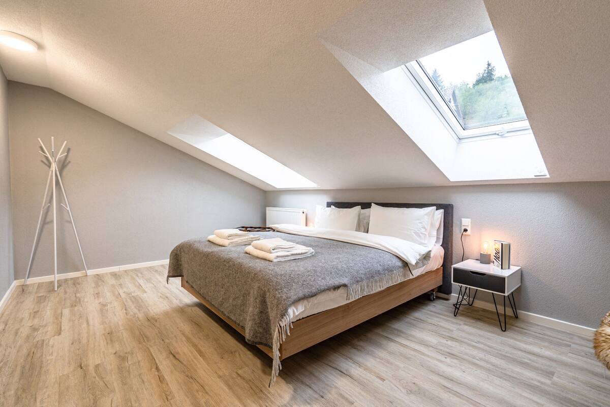 Ein modernes Schlafzimmer mit Dachfenster, Holzboden, einem Bett mit grauer Decke und einem Nachttisch mit Lampe.