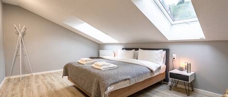 A modern bedroom with a skylight, a wooden floor, a bed with a grey blanket, and a bedside table with a lamp.