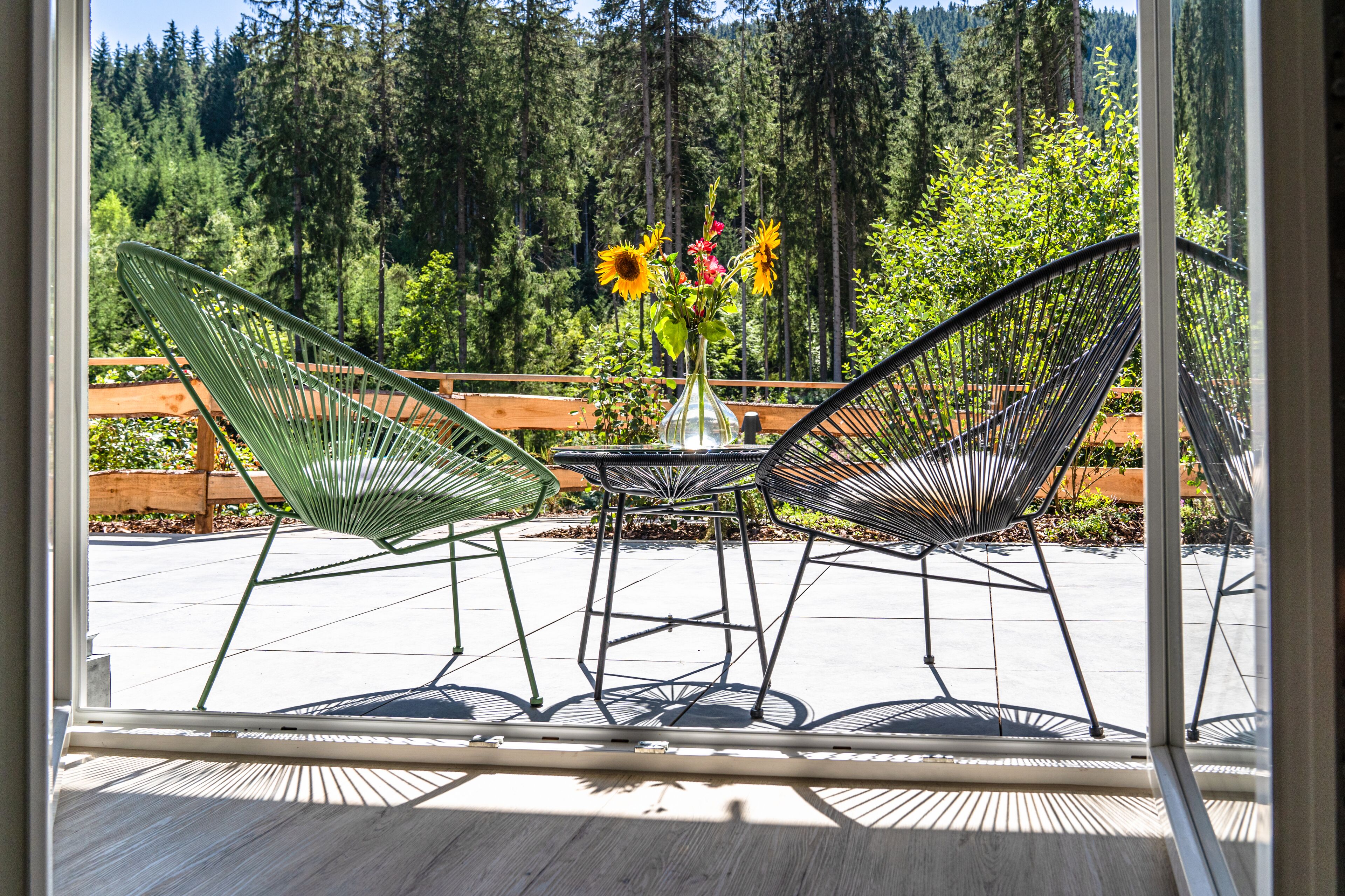 A patio with two green wire mesh chairs, a small round table, and a vase with flowers.