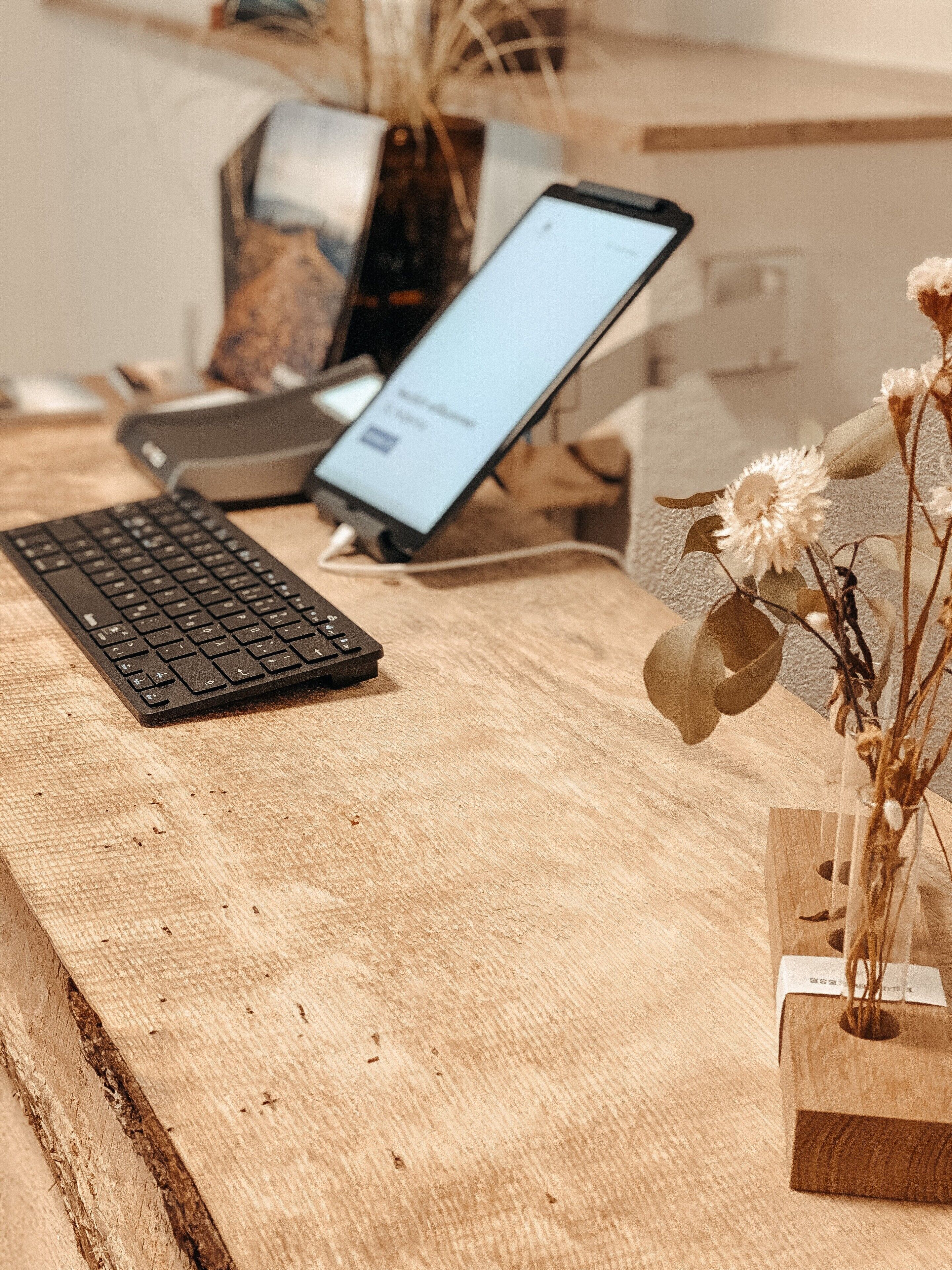 Un escritorio de madera con un teclado, una computadora portátil y un florero con flores secas.