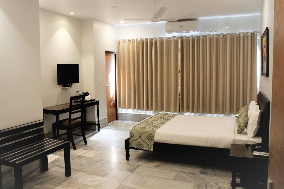 A hotel room with a bed, a desk, a chair, a bench, a television, and a window with curtains.