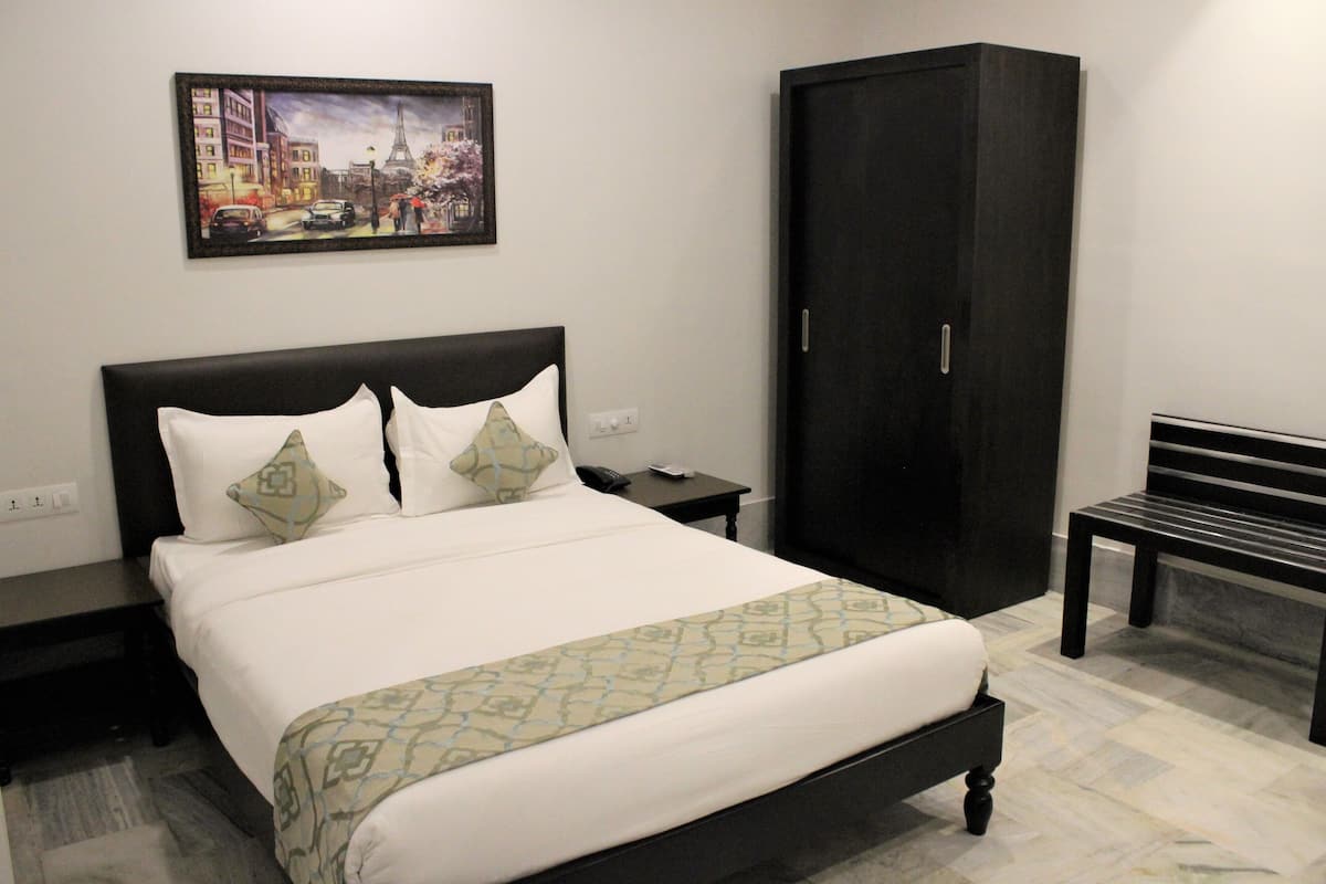A neatly made bed with a patterned bedspread, a dark headboard, and two pillows. There is a nightstand with a remote control and a phone, a dark wardrobe, and a bench in the corner.
