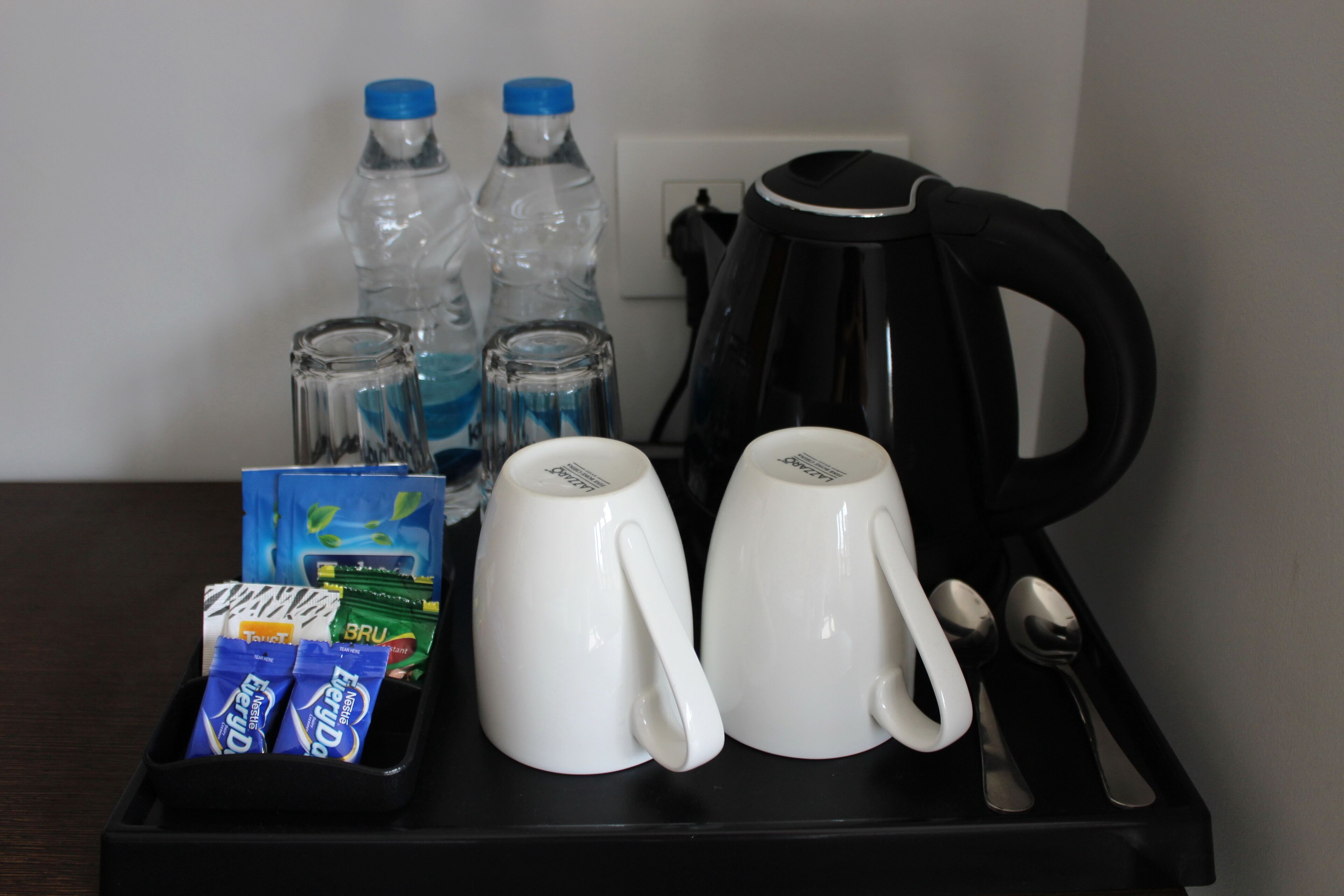 A tray with two white mugs, a black coffee maker, two water bottles, and a container with two packs of Brulee tea and a pack of Tazo tea.