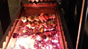 A food preparation area with skewers being grilled over an open flame, surrounded by cooking utensils and a chef in a white uniform. - Central Hotel (Phuntsholing)