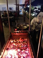 A food preparation area with skewers being grilled over an open flame, surrounded by cooking utensils and a chef in a white uniform.