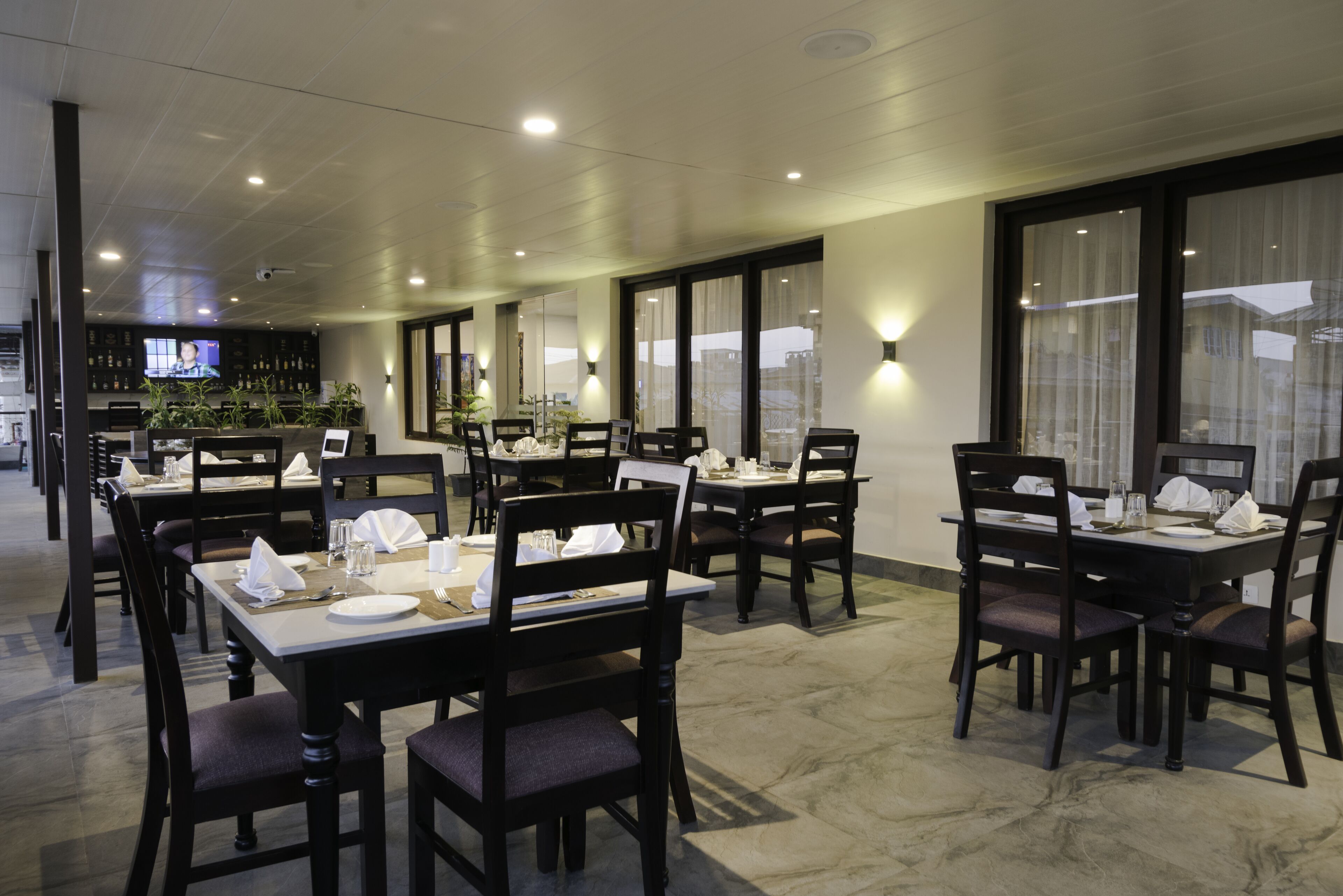 A restaurant with multiple tables and chairs, a television screen in the background, and large windows with curtains.