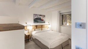 Ein Schlafzimmer mit Bett, Nachttisch, Wandlampe, Gemälde und Fenster mit Blick auf Bäume.
