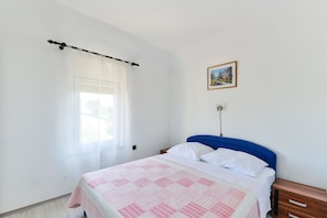 A bedroom with a bed, a nightstand, a wooden dresser, a window with sheer curtains, and a framed picture on the wall.