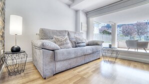 Een moderne woonkamer met een grijze bank, twee bijzettafeltjes, een vloerlamp en een groot raam met uitzicht op gebouwen en bomen.