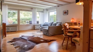 Uma sala de estar ampla com sofá, mesa de jantar com cadeiras, piso de madeira e janelas grandes com cortinas.