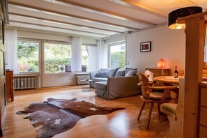 Uma sala de estar ampla com sofá, mesa de jantar com cadeiras, piso de madeira e janelas grandes com cortinas.
