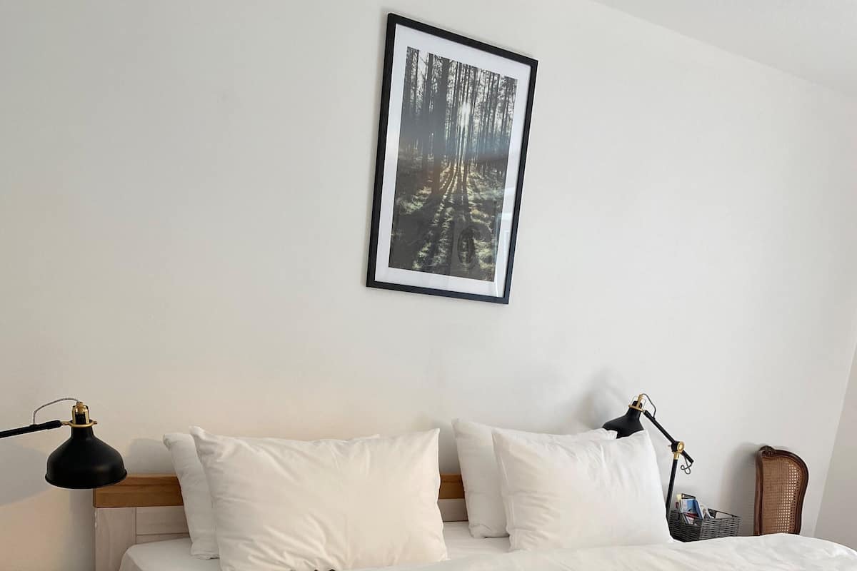 A neatly made bed with white bedding and a grey throw, a wooden nightstand with a lamp, a wicker chair, and a framed picture on the wall.