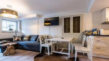 A modern living room with a sofa, a coffee table, a dining table with chairs, a flat-screen TV mounted on the wall, and a kitchen area with cabinets and a countertop.