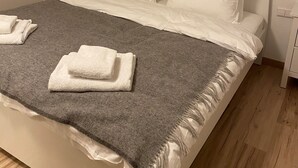 Un lit bien fait, avec du linge de lit blanc, un plaid gris et des serviettes pliées. Il y a deux lampes de chevet avec des abat-jour en bois. Un tableau encadré est accroché au-dessus du lit.