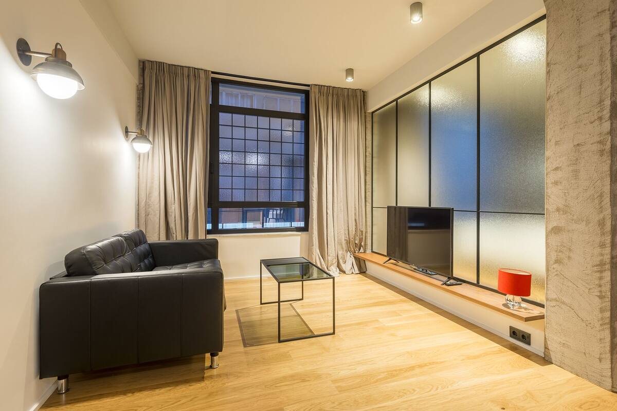 A modern living room with a black sofa, a glass coffee table, a TV, and a large window with curtains.