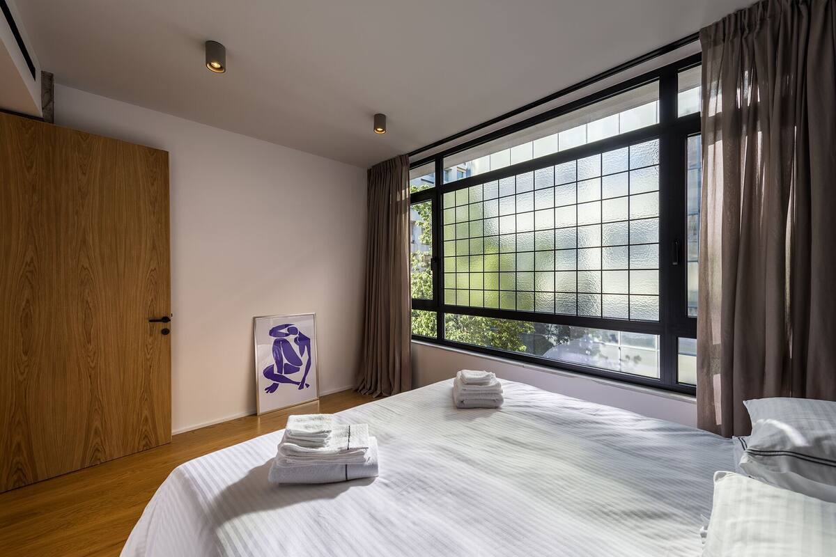 A modern bedroom with a large window, a wooden wardrobe, and a framed artwork on the wall.