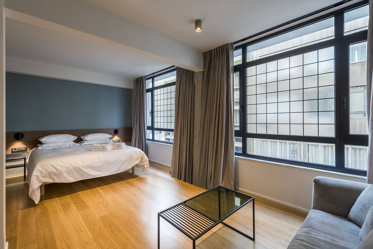 A modern hotel room with a bed, a glass coffee table, a sofa, and large windows.