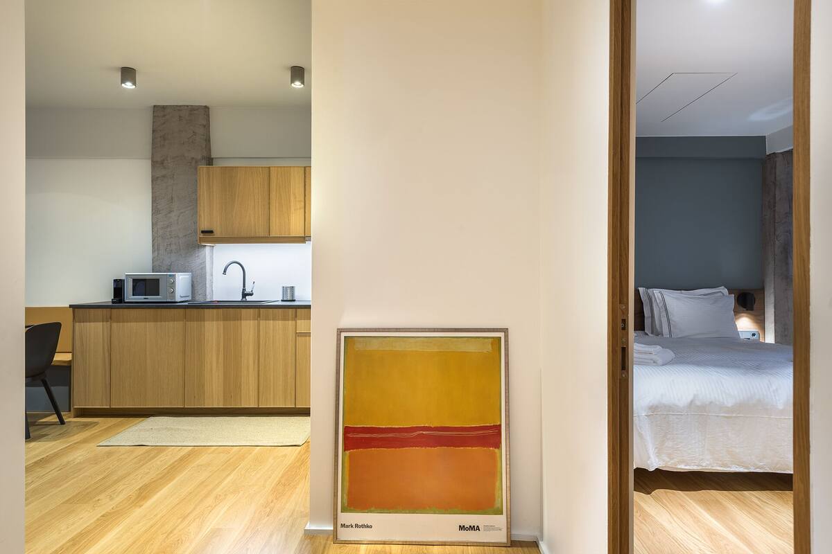 A modern hotel room with a kitchenette, a large painting, and a neatly made bed.